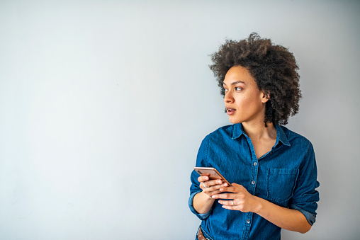 Beautiful smiling curly-haired businesswoman using smarphone. Content trendy cheerful nice cute adorable lovely girl with wavy hair in casual denim shirt, typing in phone, isolated over grey background