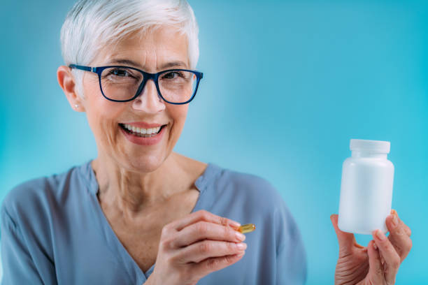 istock Supplements. Senior woman holding capsule and a blank white supplement container. 1334260035