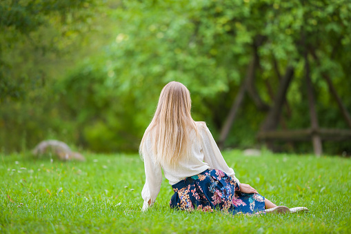 Young adult blond woman in white blouse and blue skirt sitting on green grass at natural park in summer day. Spending time alone in nature. Peaceful atmosphere. Back view. Closeup.