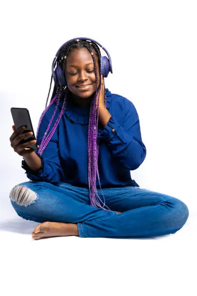 A smiling African Teenager with closed eyes listening to music via wireless headphones connected to a smartphone while squatting