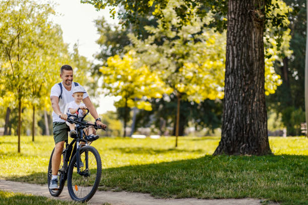 un día de familia activo en la naturaleza. un padre y su hijo andan en bicicleta por el bosque en un soleado día de verano. un chico lindo está sentado en una cesta de bicicletas mientras monta en bicicleta. disfrutan del viaje - action family photograph fathers day fotografías e imágenes de stock