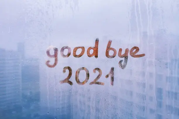 Handwritten lettering good bye and orange arabic numerals 2021 on misted glass on blue window flooded with raindrops on sunny background