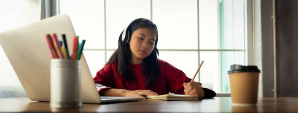istock Female student wears headphones sitting at table to learning and note, using laptop for homework during covid19 pandemic and lock down, Social Distance, New life of Student on internet. Banner Size 1334275300