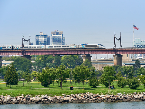 New York, NY - July 27, 2021: The Amtrak Acela Express heads north on elevated tracks over Randall's Island park in in New York City, with the East River in the foreground. Acela trains are the fastest trainsets in the Americas, attaining speeds of 150 mph.