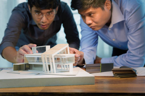istock Two asian architects discussing with designer or civil engineer about concept of building design and construction project for modern box house. Focusing on the house model. 1334280284