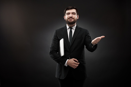 Portrait on black background in studio of businessman, holding laptop under arm, gesturing