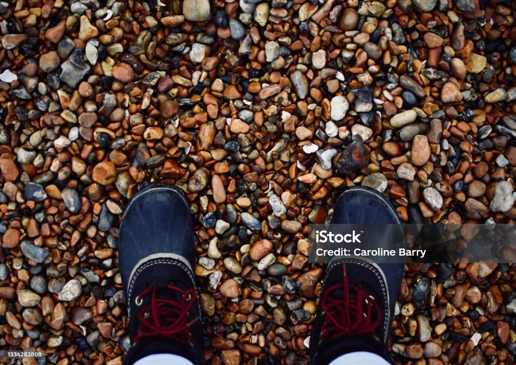 Point-of-view looking over rubber boots on a rocky beach - Brighton, UK A point of view angle of a person wearing a pair of black rubber boots with red laces and light color pants on the cold pebble beach of Brighton, England. Human Foot Stock Photo