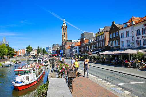 Roermond, Netherlands - July 9. 2021: View on pedestrian street with outdoor cafes and restaurants at water canal with boats, cityscape and old church tower against blue summer sky background