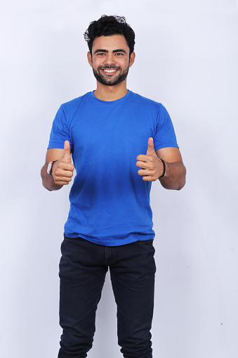 Portrait of handsome young man showing double thumbs up on white background. He is wearing blue t-shirt. Male is smiling.