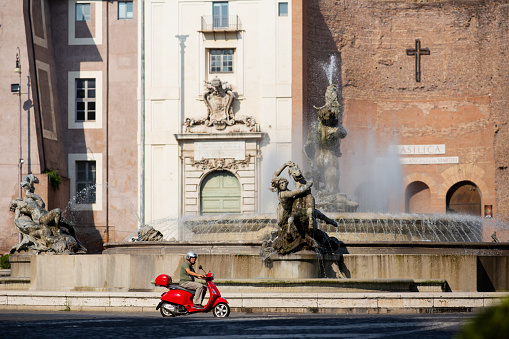 Rome, Italy - August 22, 2018: Man riding a moped through the old streets and fountain.