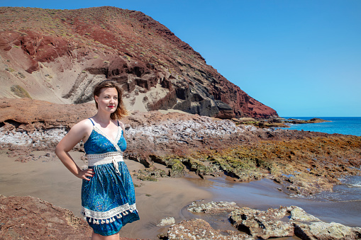 Mid adult Caucasian woman wearing a blue and white paisley dress relaxing at the rocky beach nearby Montana Roja, solo traveler in a tropical summer holidays destination in Tenerife, Canary Islands, Spain.