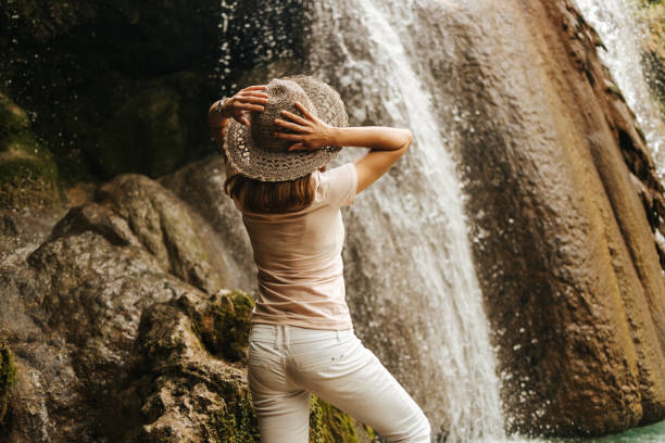 istock Beautiful young woman with a hat on her head standing next to a waterfall 1334299946