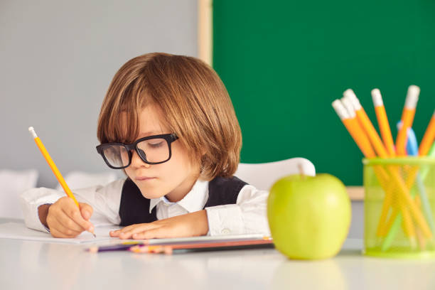 istock Back to school. Schoolboy writes with a pencil while sitting at a table against the background of the school blackboard in the classroom. 1334305481