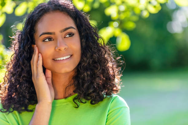 Outdoor portrait of beautiful happy mixed race African American girl teenager female young woman thinking and smiling with perfect teeth with a natural green leaves background stock photo