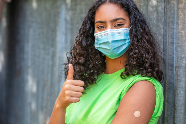 geimpfte mädchen teenager teenager gemischte rasse birassisch afroamerikanische frau junge frau mit gesichtsmaske in coronavirus covid-19-pandemie zeigt impfstoff pflaster gibt daumen hoch - thumbs up african descent teenager women stock-fotos und bilder