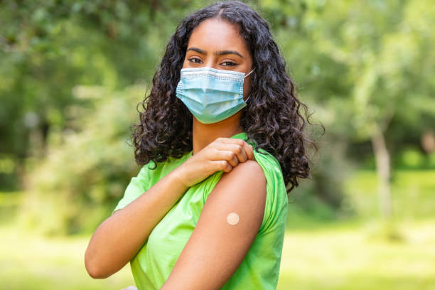 Vaccinated girl teenager teen mixed race biracial African American female young woman wearing face mask in Coronavirus COVID-19 pandemic showing vaccine band aid stock photo