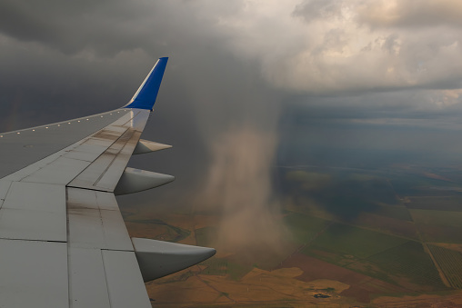 View from the window of a passenger plane on a part of the wing and pouring rain from a cloud against the background of elevated fields