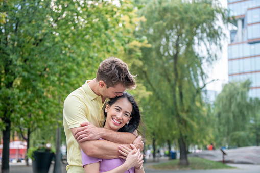 A portrait of a young and sweet heterosexual couple embracing in the city park. The boyfriend is hugging his girlfriend from behind and is kissing the top of her head. The young female adult is smiling widely at the camera.