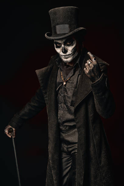 Young man in image of Baron Samedi, the Voodoo deity. Baron Saturday in black coat and top hat with stick in hand. Day of the Dead (and Halloween) theme A young man in image of Baron Samedi, the Voodoo deity. Baron Saturday in black coat and top hat with stick in hand. Day of the Dead (and Halloween) theme face paint halloween adult men stock pictures, royalty-free photos & images