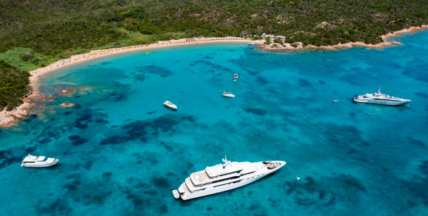 View from above, stunning aerial view of a green coastline with a white sand beach and and boats sailing on a turquoise water at sunset. Cala di volpe beach, Costa Smeralda, Sardinia, Italy. View from above, stunning aerial view of a green coastline with a white sand beach and and boats sailing on a turquoise water at sunset. Cala di volpe beach, Costa Smeralda, Sardinia, Italy. Cala Di Volpe stock pictures, royalty-free photos & images