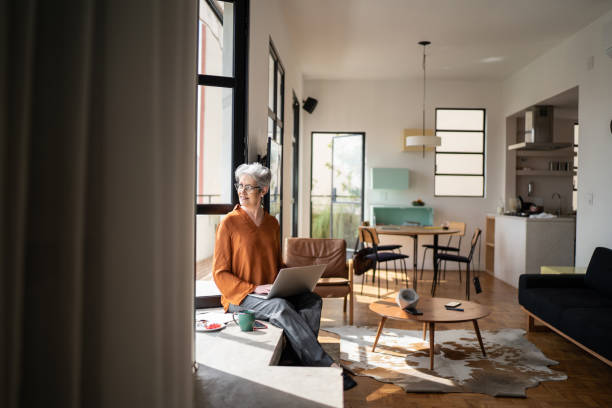 mujer mayor contemplando y usando la computadora portátil en casa - full length windows fotografías e imágenes de stock