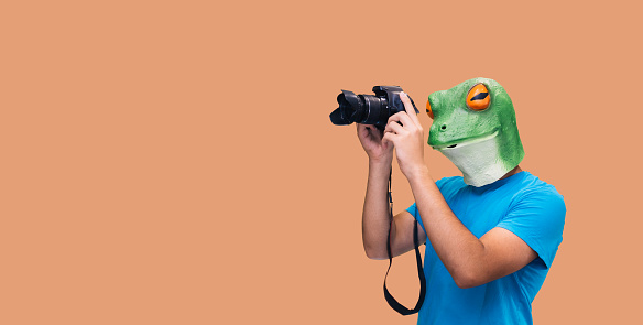 person in frog mask gesture with a camera as a photojournalist looking through the lens on orange background with copy space