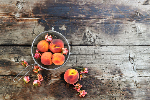 Ripe peaches in a sita with dried rose flower buds, on a textured wooden background.