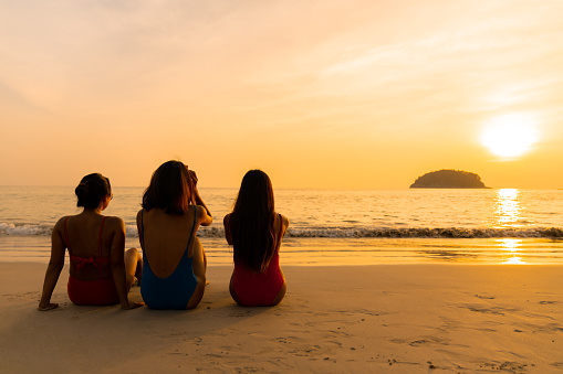 Group of Confidence Asian woman friendship in colorful swimwear sitting on the beach and talking together at summer sunset. Female girl friends enjoy and having fun outdoor activity lifestyle on summer vacation.