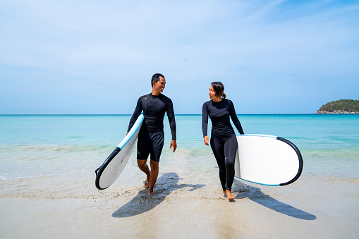 Happy Asian family enjoy outdoor active lifestyle play extreme sports surfing on summer vacation. Healthy couple in wetsuit holding surfboard and walking together on the beach at summer sunny day.