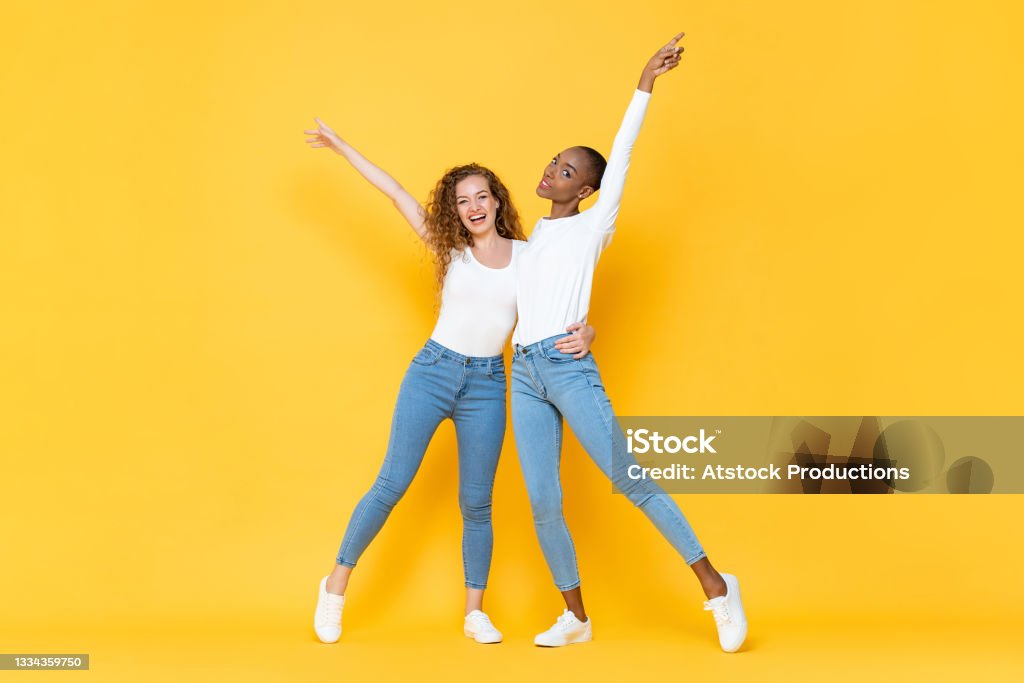 Full length portrait of smiling two interracial millennial woman friends holding each other and raising hands up in isolated studio yellow color background 30-34 Years Stock Photo