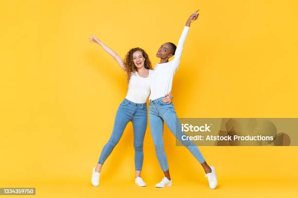 Full Length Portrait Of Smiling Two Interracial Millennial Woman Friends Holding Each Other And Raising Hands Up In Isolated Studio Yellow Color Background Stock Photo - Download Image Now