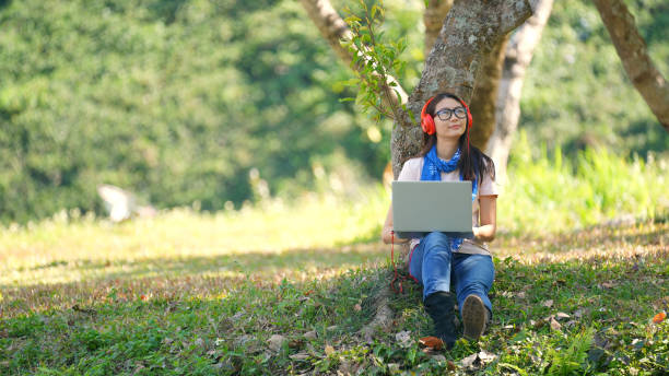 공원에서 헤드폰에서 음악을 듣고 노트북에서 일하는 젊은 아시아 여성 - laptop computer grass nature 뉴스 사진 이미지