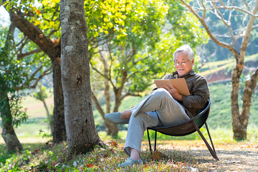 Asian senior man sitting alone on outdoor chair in nature park with writing or drawing on paper notebook. Healthy elderly retired male relax and enjoy outdoor leisure activity hobby in sunny day