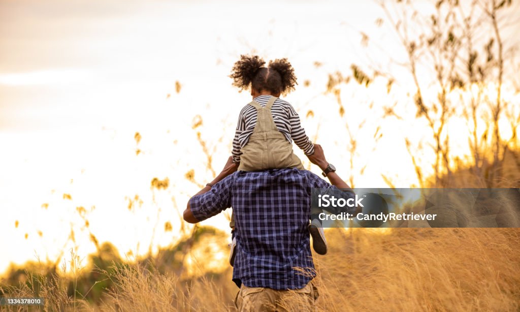 Mixed race family hiking together on the meadow at summer sunset Happy mixed race family on holiday vacation. Smiling father and little daughter carrying and hiking together on meadow hill in summer. Parent with cute child girl relax and enjoy outdoors lifestyle. Family Stock Photo