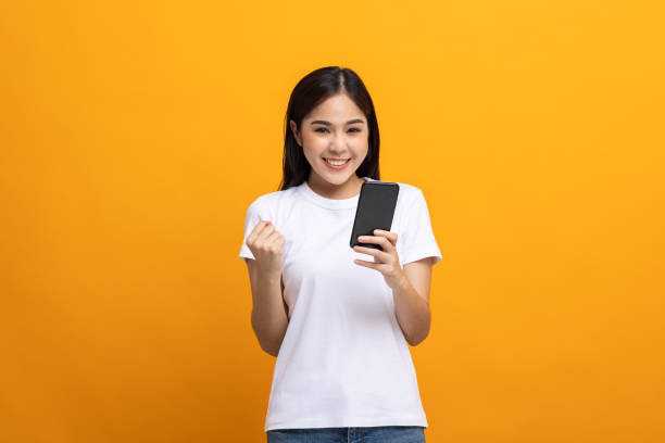 istock Pretty young asian woman using smartphone standing on isolated yellow background feeling happy. Shopping online payment with mobile phone. Excited and surprised female holding cellphone. 1334404518