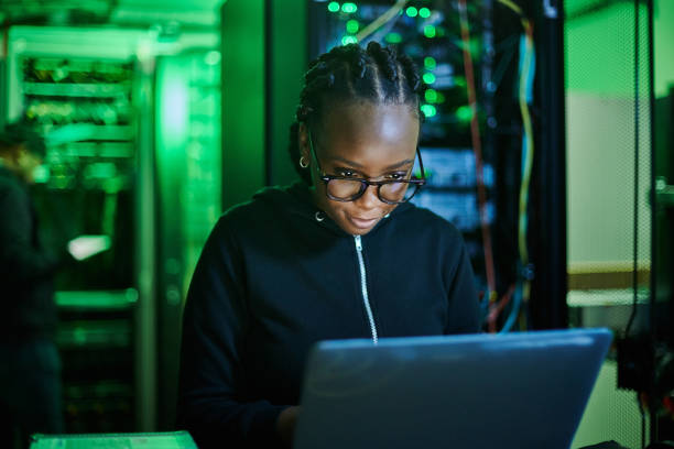 Shot of a young female computer programer using a laptop while standing in a dark server room Hackers describe what they do as creative problem solving. black nerd stock pictures, royalty-free photos & images