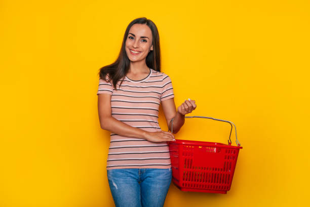 Beautiful young happy woman customer with red grocery basket is posing on yellow background Beautiful young happy woman customer with red grocery basket is posing on yellow background holding shopping basket stock pictures, royalty-free photos & images