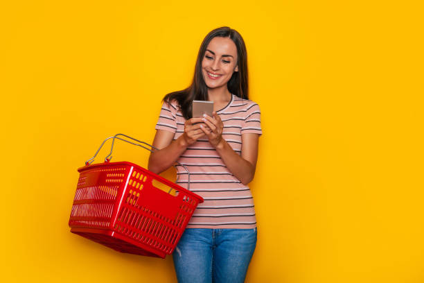 Beautiful young happy woman customer with red grocery basket is posing on yellow background Beautiful young happy woman customer with red grocery basket is posing on yellow background holding shopping basket stock pictures, royalty-free photos & images