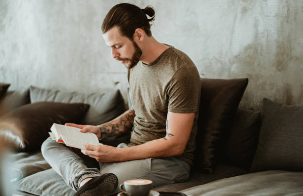 porträt eines jungen bärtigen hipsters, der im café sitzt, während er bücher liest und zur seite schaut. - shirt paper book business stock-fotos und bilder