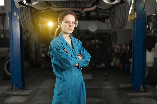 A young woman in work clothes, an apprentice stands next to the car and is proud and happy in the garage with protective goggles. Copy space.