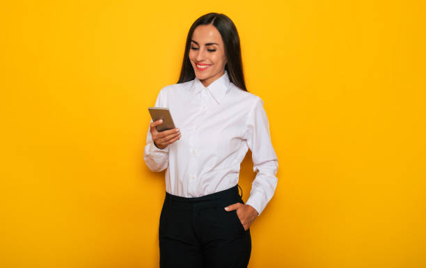 Modern happy successful confident business woman in a white shirt with smart phone is posing on yellow background stock photo