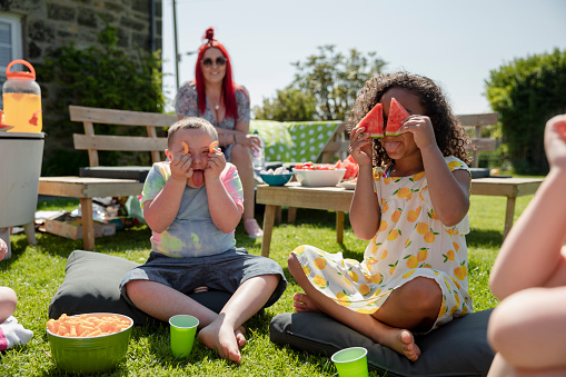 Children sitting outside having a picnic together in summer in the North East of England. Some of them are holding fruit up to their faces sticking out their tongues.