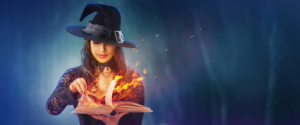 Halloween Witch girl with magic Book of spells portrait. Beautiful young woman in witches hat conjuring, making witchcraft. Over spooky dark magic forest background. Wide Halloween party art design. stock photo