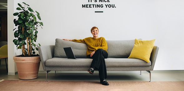 Smiling businesswoman sitting on sofa in office lobby. Cheerful entrepreneur on sofa.