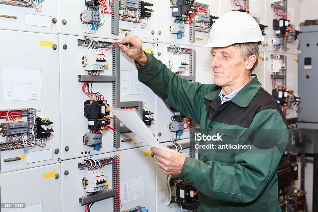 Senior worker standing near electrical panel with an electric screwdriver Electrician standing near electrical shield with an electric screwdriver. White helmet Construction Site Stock Photo