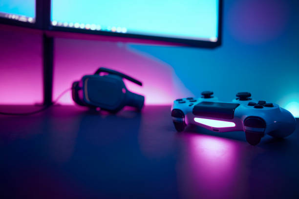 istock Gaming controller laying on dark table in front of dual lcd display. Light blue and pink illuminated wall in background. 1334436134