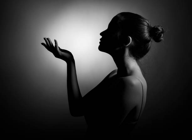 A silhouette of a beautiful young woman in profile with her palm outstretched against a dark background. stock photo