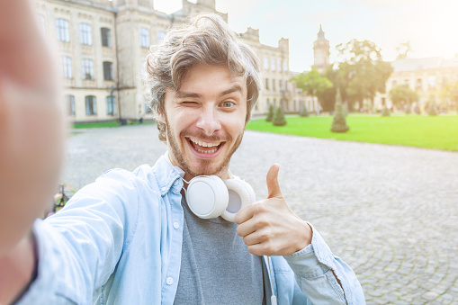 Portrait of happy young man is making selfie photo with thumb up on background of university campus. Smiling student is walking outdoor. Ready to study. Education concept. Back to high school.