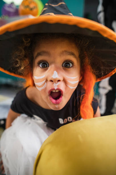 Boo!!! A close-up front view of a little girl with curly hair and brown eyes looking directly into camera and pulling a funny face. She is dressed in a witches hat with a wig inside of it and is all dressed and ready for Halloween and trick-or-treating. halloween face paint stock pictures, royalty-free photos & images