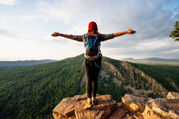 A traveler girl with a backpack is standing on the edge of the mountain, a rear view. A young woman with a backpack standing on the edge of a cliff and looking at the sky with her hands raised. stock photo